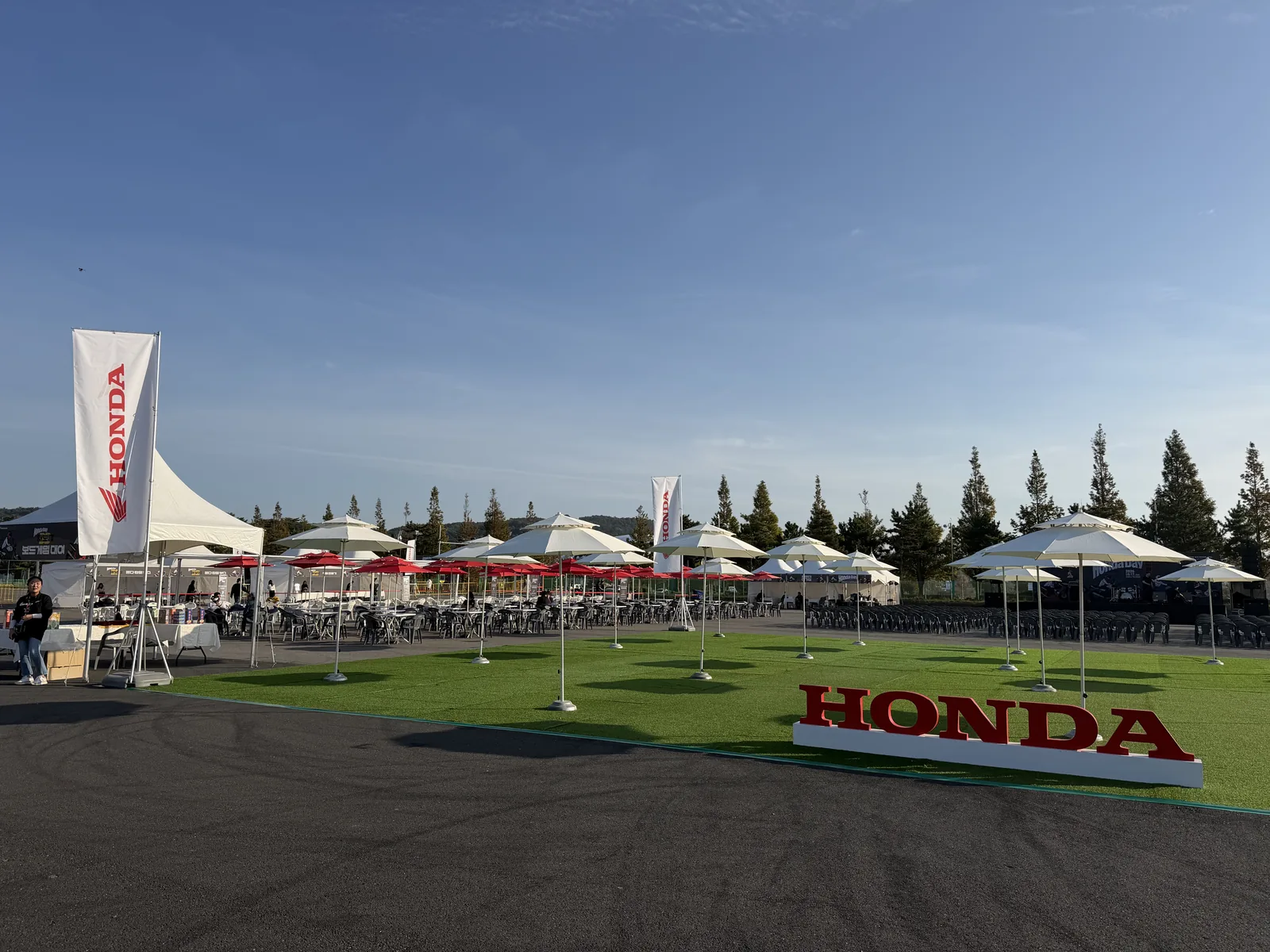 광장을 통째로 쓴 제11회 혼다데이 터 — 가운데 빨간 입체 HONDA 글자, 윙 로고를 넣은 세로 깃발 둘, 펴 놓은 파라솔 밑으로 늘어선 수백 석의 앉을 자리, 한쪽 끝의 HONDA DAY 백월과 흰 천막, 등 뒤편의 야산과 침엽수