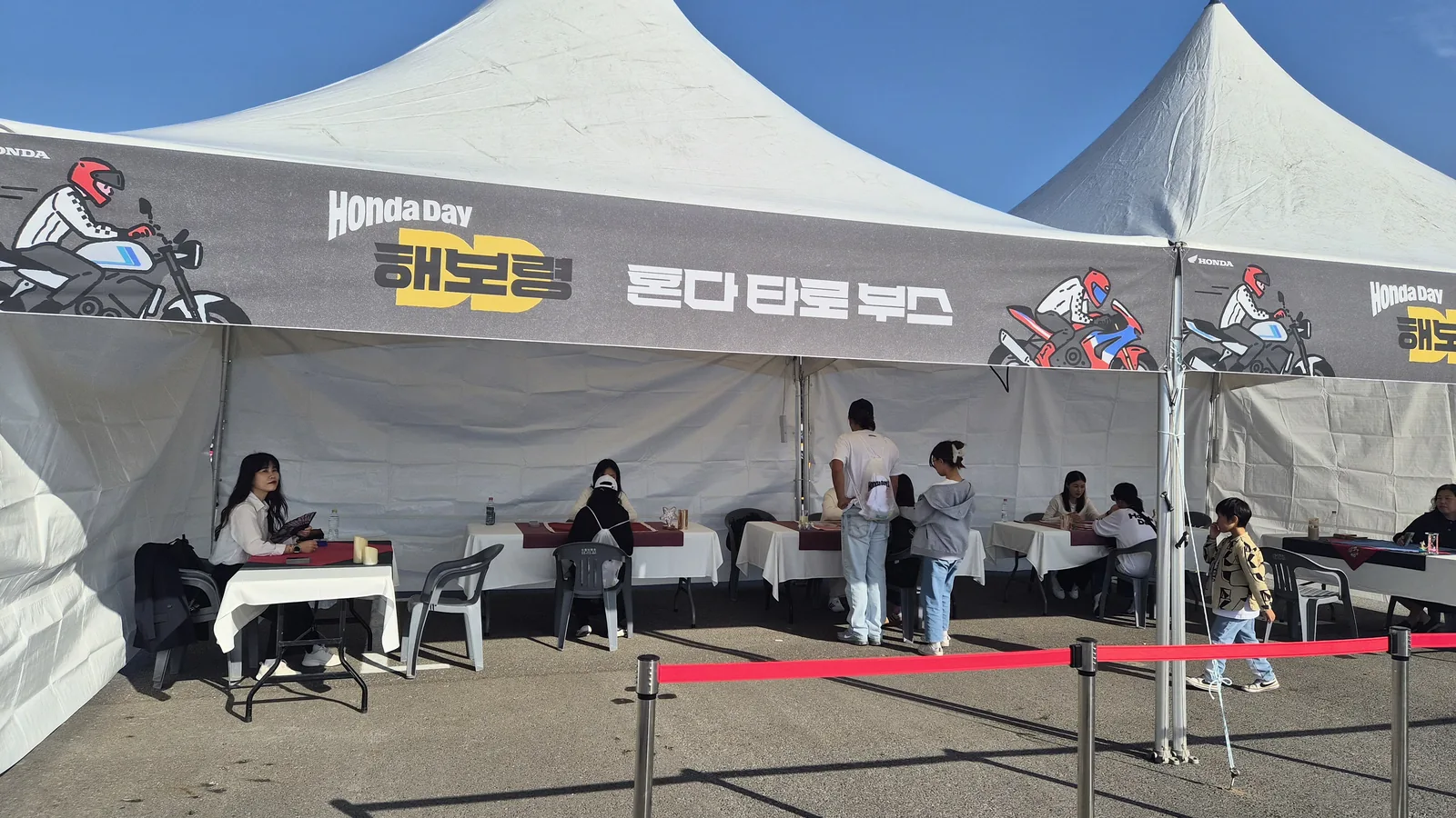 한 줄로 늘어선 타로 상담 자리 다섯을 천막 정면에서 본 모습 — 흰 테이블보 위에 와인색 타로천을 깔고 타로카드를 올려 두었으며, 위쪽 처마로 HONDA 윙 로고와 「혼다 타로 부스」 배너가 보인다