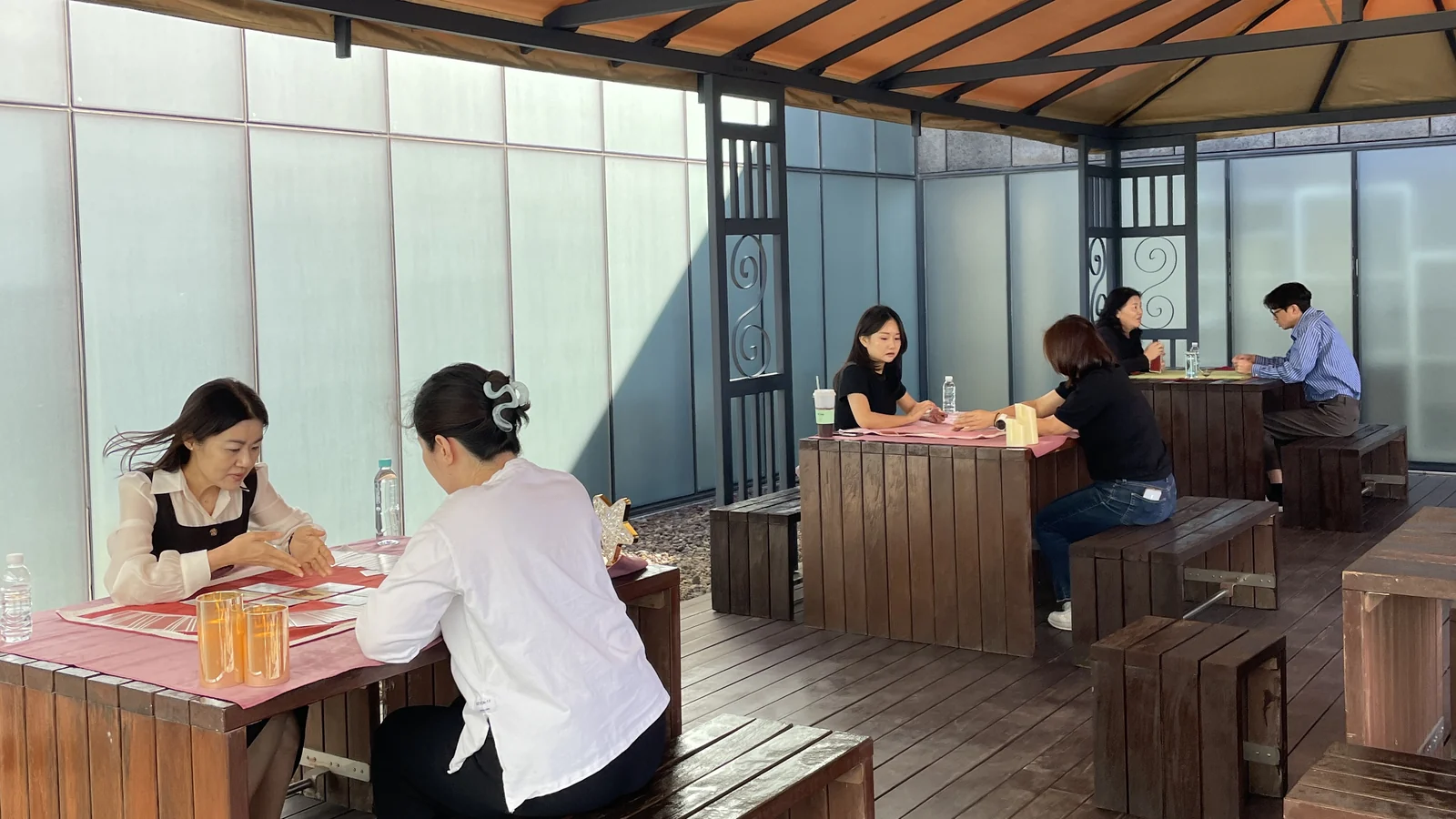 주황 천막을 덮은 정자 아래 목재 테이블 세 자리에서 타로 상담이 함께 진행되는 교보문고 본사 테라스 — 붉은·분홍 매트 위에 펼쳐진 카드와 테이블마다 놓인 음료·생수
