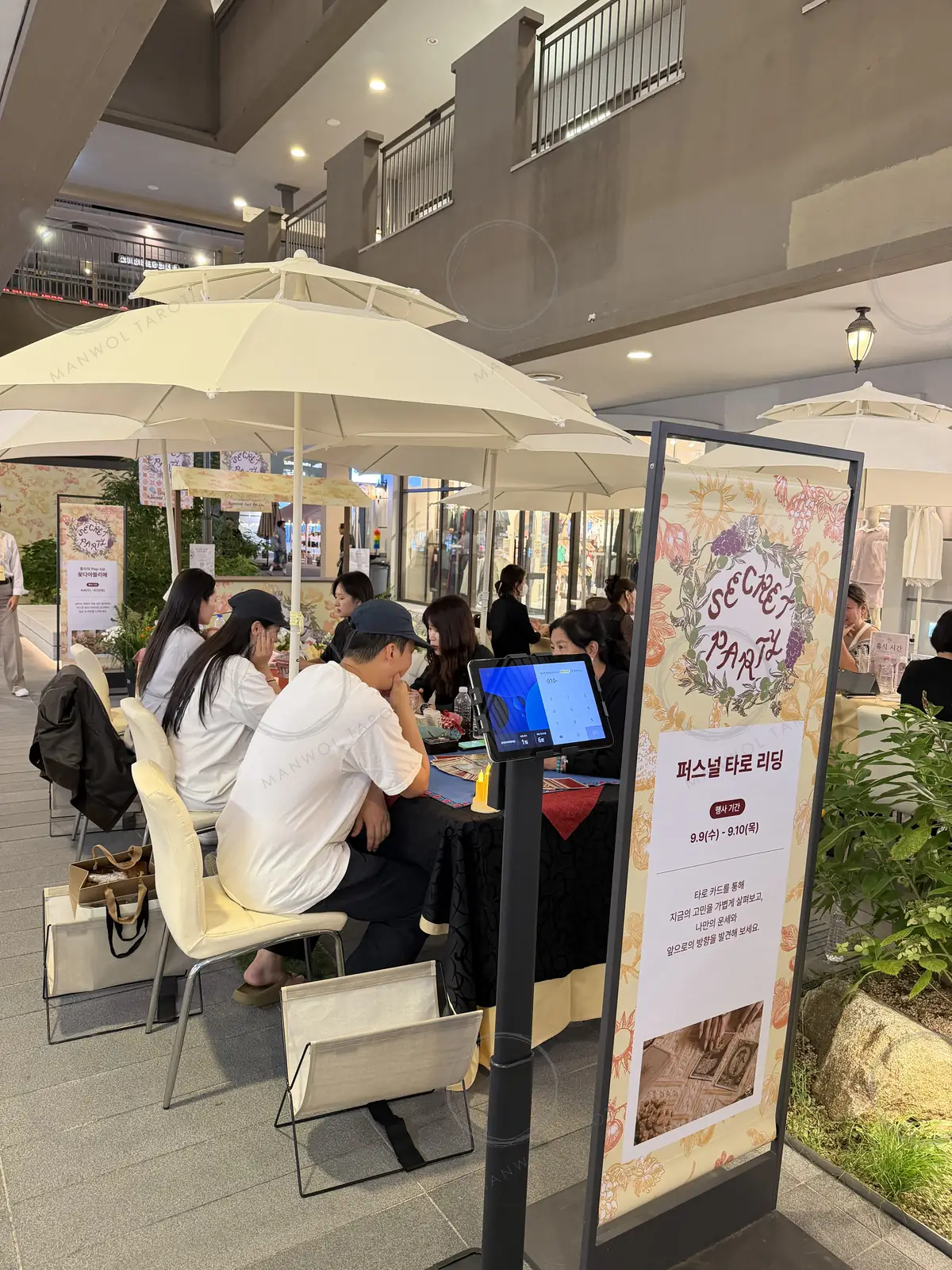 아울렛 통로의 흰 파라솔 아래 타로 상담 테이블 — 고객들이 전문가와 마주 앉아 카드를 보고 있고, 앞쪽에 「퍼스널 타로 리딩」 안내 배너가 서 있다
