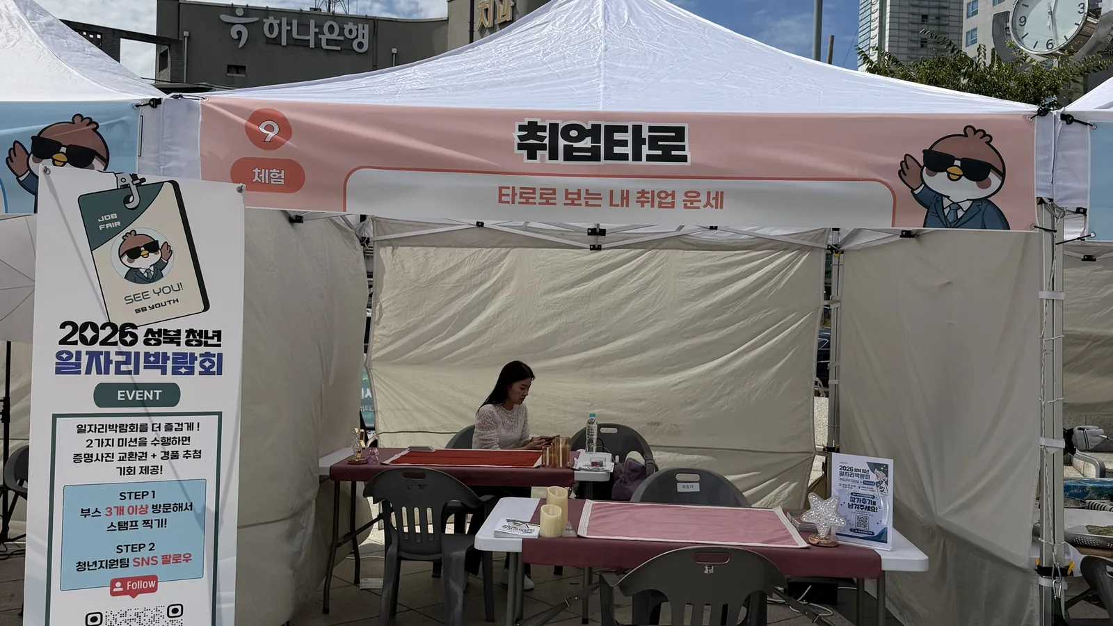 분홍 현수막을 앞면에 건 흰 차양 아래 차려진 타로 상담 자리 — 붉은 매트를 깐 상판 두 자리와 초 모양·별 모양 장식 소품
